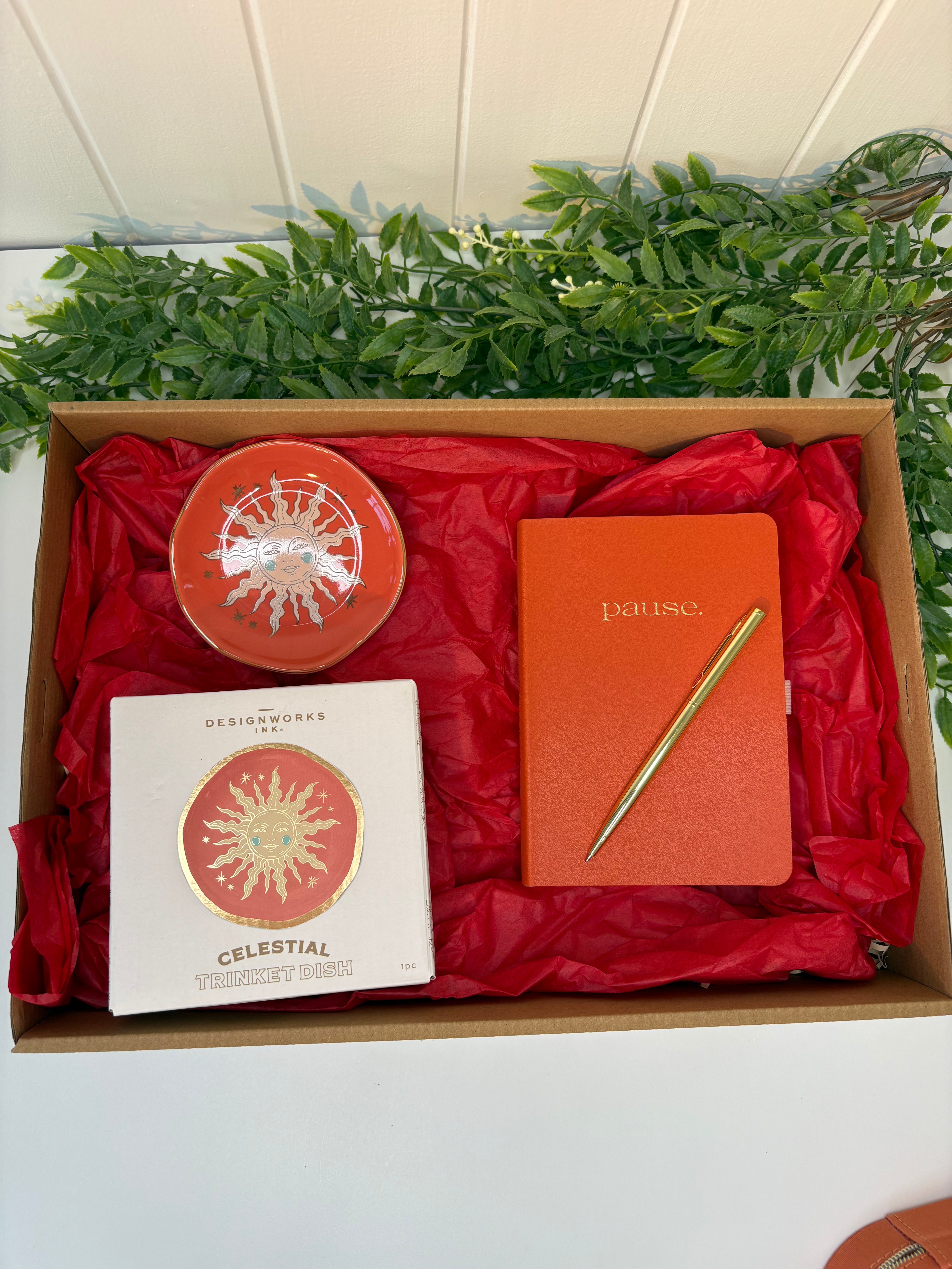 Gift box with red interior containing an orange notebook, pen, and decorative items on a white surface with greenery.